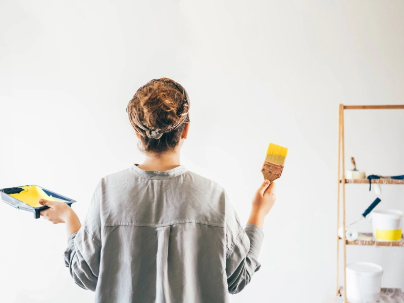 woman-with-paint-brush-dipped-in-yellow-paint-looking-at-white-wall