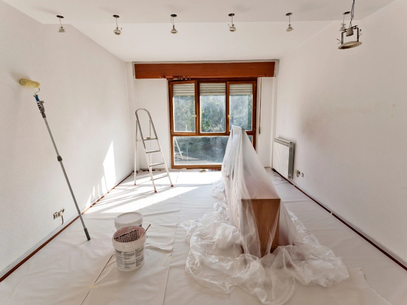 room-prepared-to-be-painted-with-clear-plastic-tarp-overfurniture-and-roller-paper-on-floor-and-roller-paint-brush-with-pole-extender-leaning-against-wall